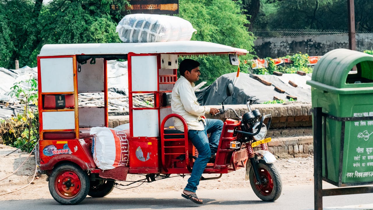A man driving a red bicycle rickshaw loaded with goods through an urban street.
